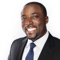 Photo of attorney Kwame Christian in a black suit and blue tie, smiling at the camera, view from chest up, on white background. 
